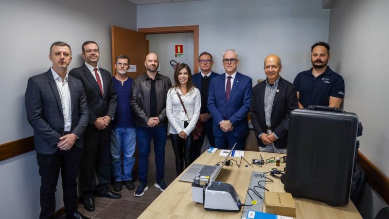 No primeiro plano, mais &agrave; direita, nateriais de laborat&oacute;rio aparecem dispostos sobre mesa de madeira. No fundo, nove pessoas, uma ao lado da outra