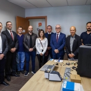 No primeiro plano, mais &agrave; direita, nateriais de laborat&oacute;rio aparecem dispostos sobre mesa de madeira. No fundo, nove pessoas, uma ao lado da outra